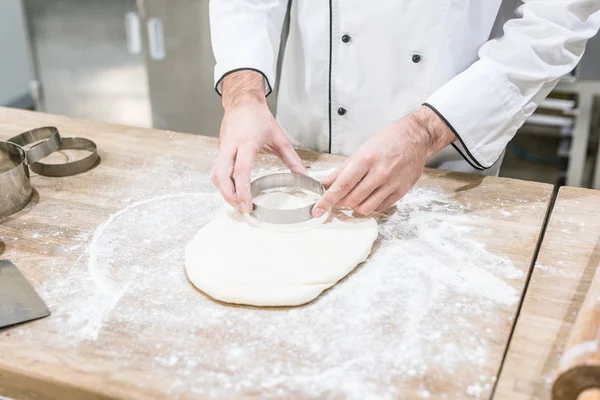 Cropped view of baker hands cutting dough with round cutter on wooden table — Stock Photo