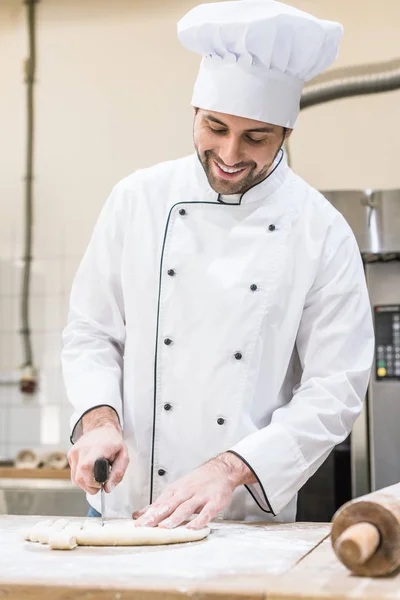 Handsome baker smiling and cutting uncooked dough on wooden table — Stock Photo