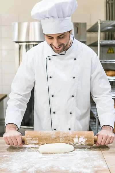 Улыбающийся пекарь катит сырое тесто на деревянный стол — стоковое фото