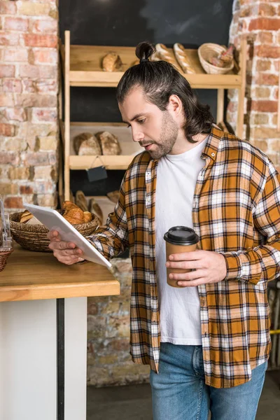 Handsome seller holding digital tablet and paper cup in bakehouse — Stock Photo