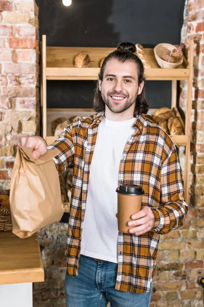 Handsome seller standing with paper cup and paper bag — Stock Photo