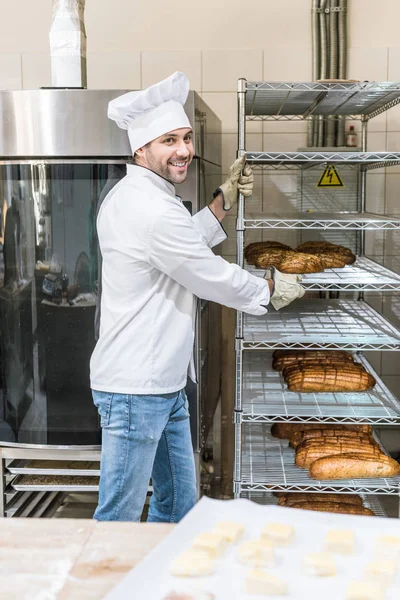 Vista lateral del chef sonriente sacando estante con pan - foto de stock
