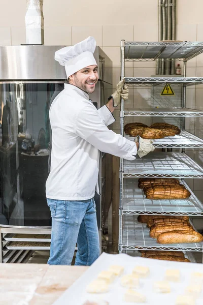 Side view of smiling chef taking out rack with bread — Stock Photo