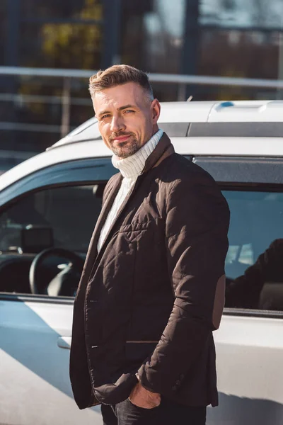 Веселый мужчина стоит и улыбается возле машины с руками в карманах — стоковое фото