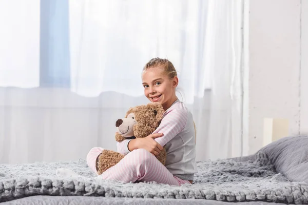 Fröhliches Kind hält Teddybär und sitzt auf Bett — Stockfoto