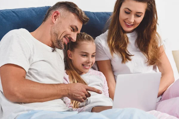 Père souriant pointant vers un ordinateur portable et couché dans la chambre avec sa femme et sa fille — Photo de stock