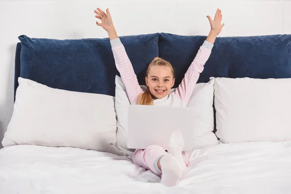 Alegre niño preadolescente acostado en la cama con el ordenador portátil y poniendo las manos arriba - foto de stock