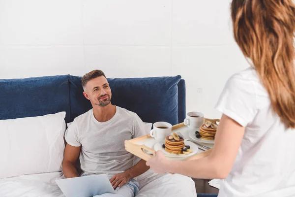 Marido sentado com laptop e olhando para a esposa segurando café da manhã na bandeja — Fotografia de Stock