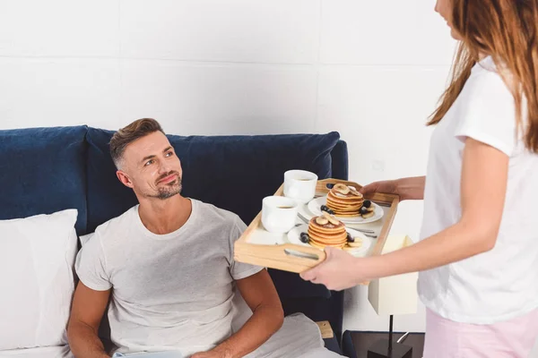 Mulher segurando bandeja com café da manhã e marido deitado na cama — Fotografia de Stock