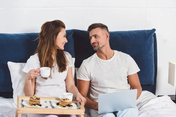 Husband looking at wife and sitting with laptop in bed — Stock Photo