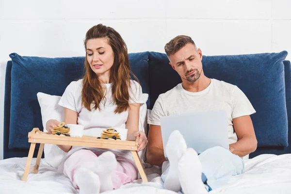 Husband using laptop and wife having breakfast in bed — Stock Photo
