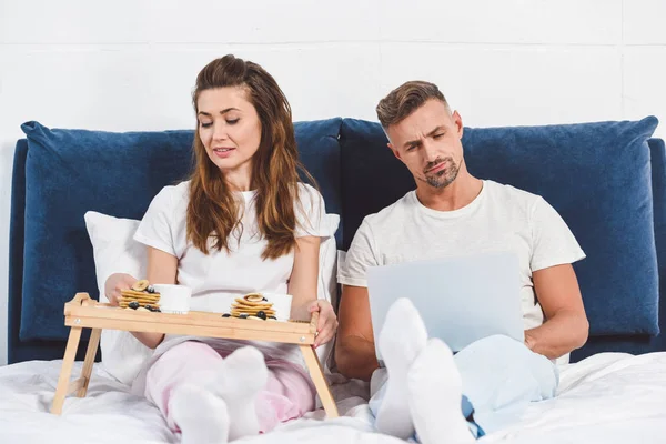 Mari utilisant ordinateur portable et femme prenant le petit déjeuner au lit — Photo de stock