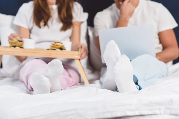 Vista recortada del marido usando el ordenador portátil, mientras que la esposa desayunando en la cama - foto de stock