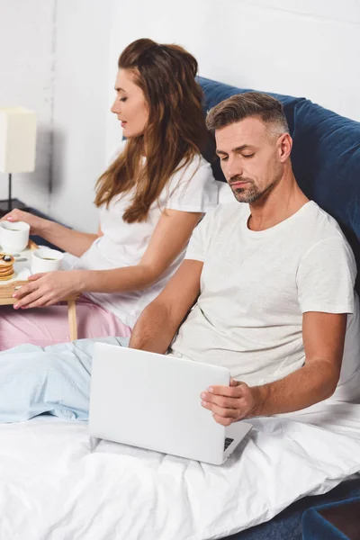 Schöner Mann benutzt Laptop, während attraktive Frau im Bett frühstückt — Stockfoto