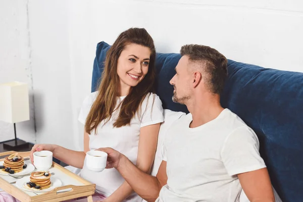 Casal alegre conversando e tomando café da manhã na cama de manhã — Fotografia de Stock