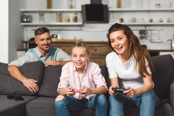 Улыбающиеся мать и дочь играют в видеоигры, пока отец прячется за диваном — стоковое фото