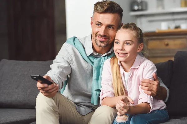 Padre con mando a distancia sentado en el sofá y abrazando a la hija - foto de stock