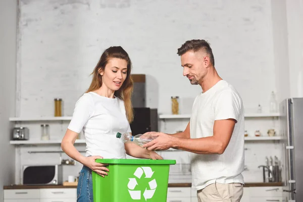 Pareja de adultos poniendo botellas de plástico vacías en la caja verde de reciclaje en la cocina - foto de stock