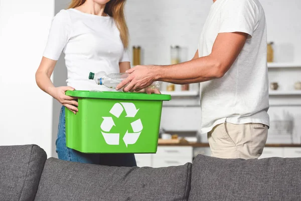 Vista recortada de pareja de adultos poniendo botellas de plástico vacías en la caja verde de reciclaje - foto de stock