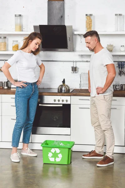 Adult couple with hands on heaps standing at kitchen and looking at green recycle box — Stock Photo