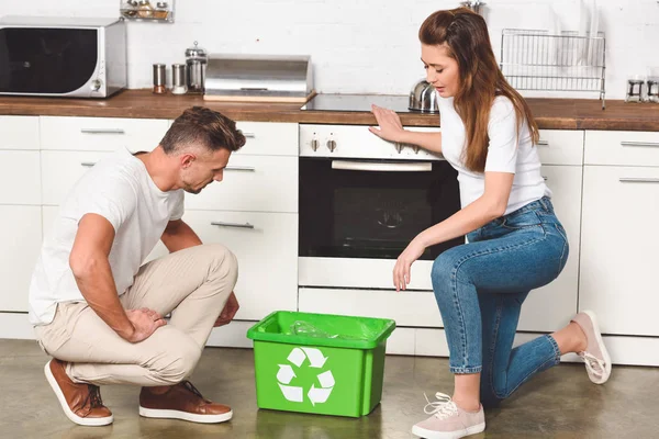 Pareja de adultos de pie en la cocina con caja de reciclaje verde en el suelo - foto de stock