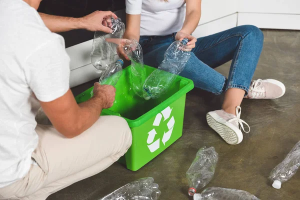 Vista recortada de la pareja sentada en el suelo y poniendo botellas de plástico en la caja verde en la cocina - foto de stock