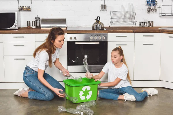 Madre e hija sentadas en el suelo en la cocina y poniendo botellas de plástico vacías en la caja con cartel de reciclaje - foto de stock