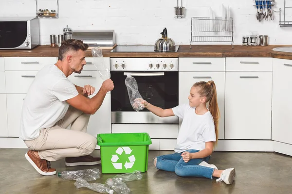 Padre e figlia seduti sul pavimento in cucina e mettere bottiglie di plastica vuote in scatola con segno di riciclo — Foto stock