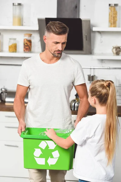 Pai e filha colocando garrafas de plástico vazias na caixa de reciclagem verde na cozinha — Fotografia de Stock