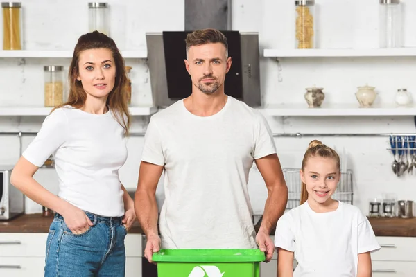 Familia de pie en la cocina con caja de reciclaje verde y mirando a la cámara juntos - foto de stock