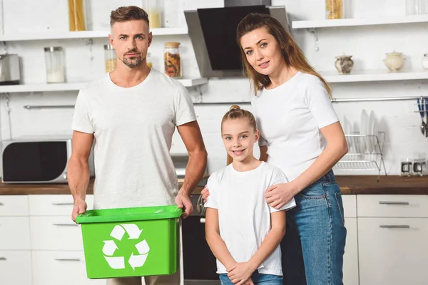 Familia de pie en la cocina con caja de reciclaje verde y mirando a la cámara - foto de stock