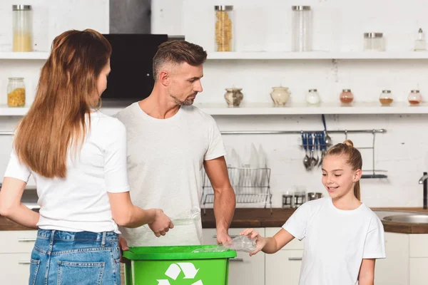 Familia de pie en la cocina y poner botellas de plástico vacías en la caja de reciclaje verde - foto de stock