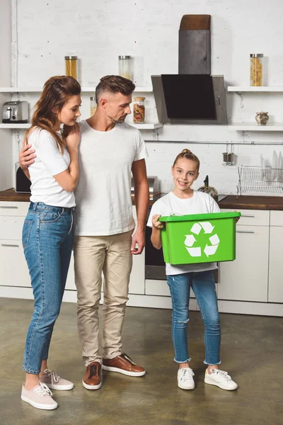 Padres de pie en la cocina mientras su hija sostiene la caja de reciclaje verde - foto de stock