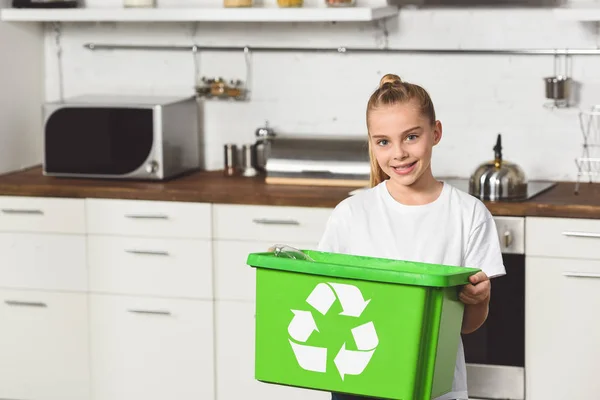 Niño sonriente de pie en la cocina con caja de reciclaje verde - foto de stock