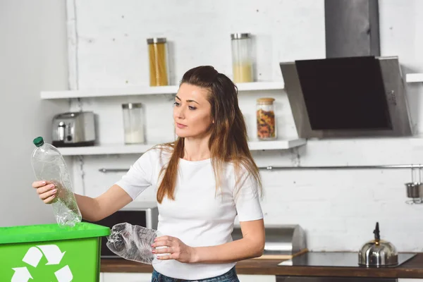 Mulher colocando garrafas de plástico em caixa de reciclagem verde na mesa de madeira na cozinha — Fotografia de Stock