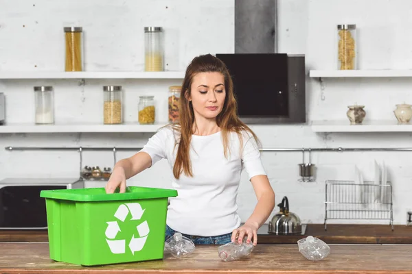 Attraktive Frau legt Plastikflaschen in grüne Recyclingbox an Holztisch — Stockfoto