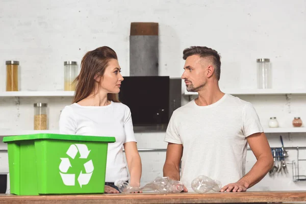 Adult couple looking at each other with green recycle box and empty plastic bottles at kitchen table — Stock Photo