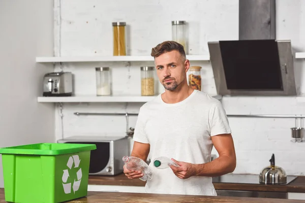 Красивый мужчина держит пластиковые бутылки с зеленой коробки утилизации за деревянным столом — стоковое фото
