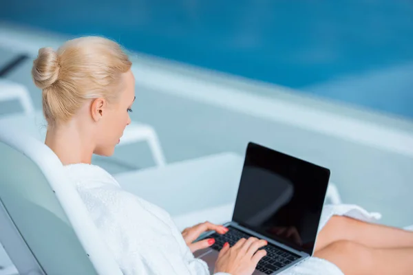 Femme utilisant un ordinateur portable en peignoir blanc près de la piscine — Photo de stock