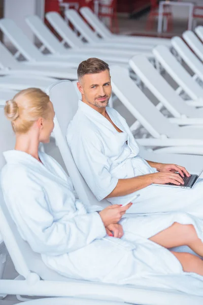 Alegre marido acostado en la cubierta de la silla con el ordenador portátil y mirando a la esposa - foto de stock