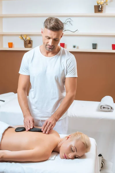 Gutaussehender Therapeut legt Frau heiße Steine auf den Rücken — Stockfoto