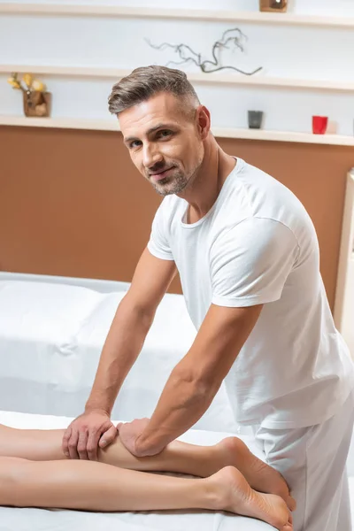 Adult handsome man massaging woman legs and looking at camera in spa — Stock Photo