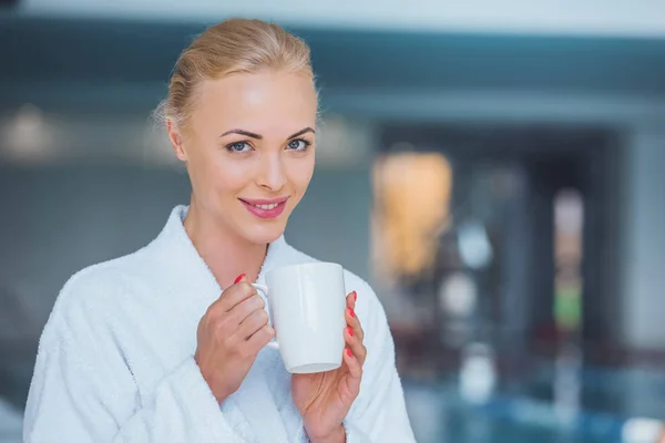 Sonriente atractiva mujer rubia sosteniendo taza blanca en spa - foto de stock