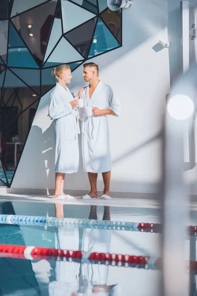 Pareja en albornoces blancos de pie cerca de la piscina con bebidas - foto de stock