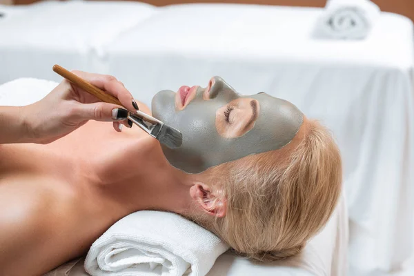 Close up de esteticista aplicando máscara de barro com escova no rosto da mulher — Fotografia de Stock
