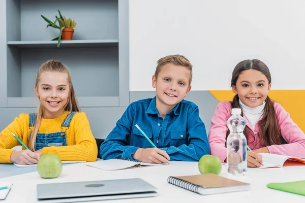 Classmates writing in notebooks and smiling at camera during lesson — Stock Photo