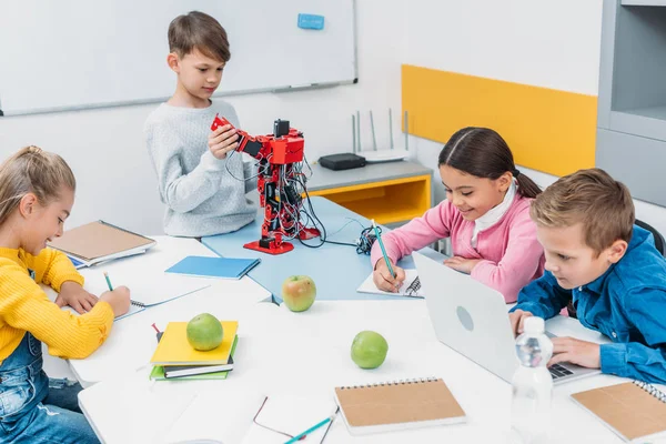 Schoolchildren writing, using laptop, working on robot at STEM robotics lesson — Stock Photo