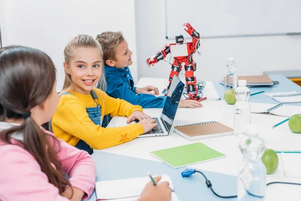 Bambini che scrivono nel taccuino e usano il computer portatile alla scrivania con robot rosso in classe stelo — Foto stock