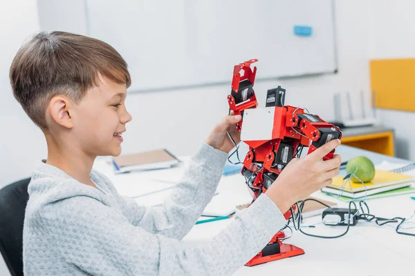 Smiling schoolboy playing with red handmade robot in classroom — Stock Photo