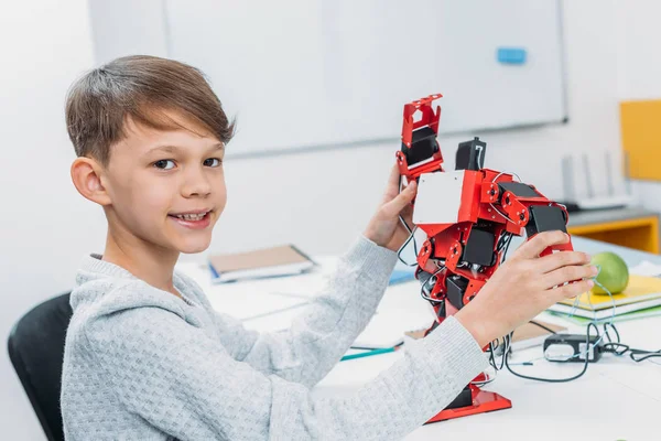 Sorridente scolaro guardando la fotocamera e tenendo il robot fatto a mano rosso in classe — Foto stock